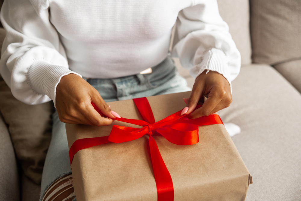 Close up hands of young woman opens box with holiday gift with red ribbon.
