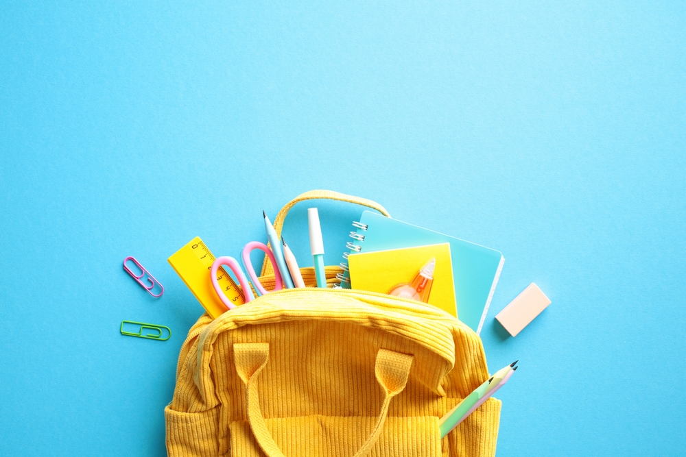 Back to School Concept. Brightly colored yellow backpack overflowing with essential school supplies such as notebooks, pens, pencils, scissors, and a ruler on a vibrant blue background. Flat lay.