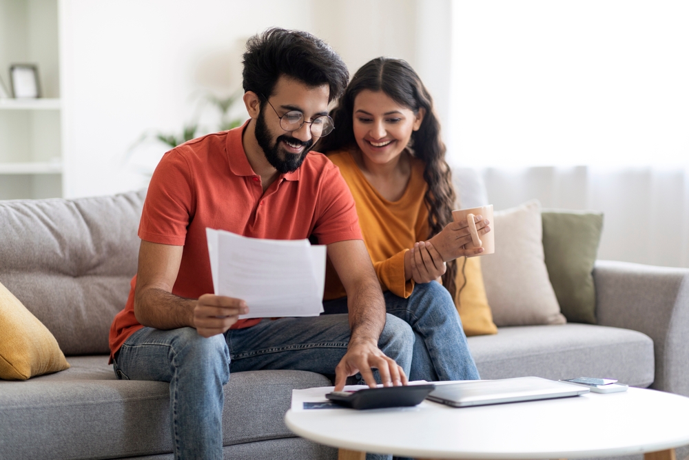 Happy Indian Spouses Checking Financial Documents And Calculating Family Budget At Home, Cheerful Eastern Couple Sitting At Table In Living Room, Reading Loan Papers Or Counting Monthly Expenses
